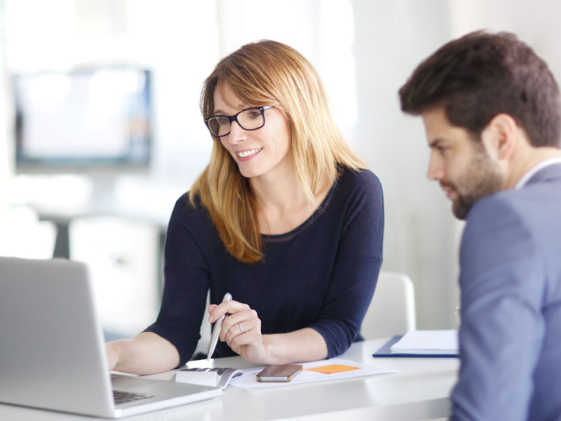 Portrait of investment advisor businesswoman sitting at office in front of computer and consulting with young professional man.